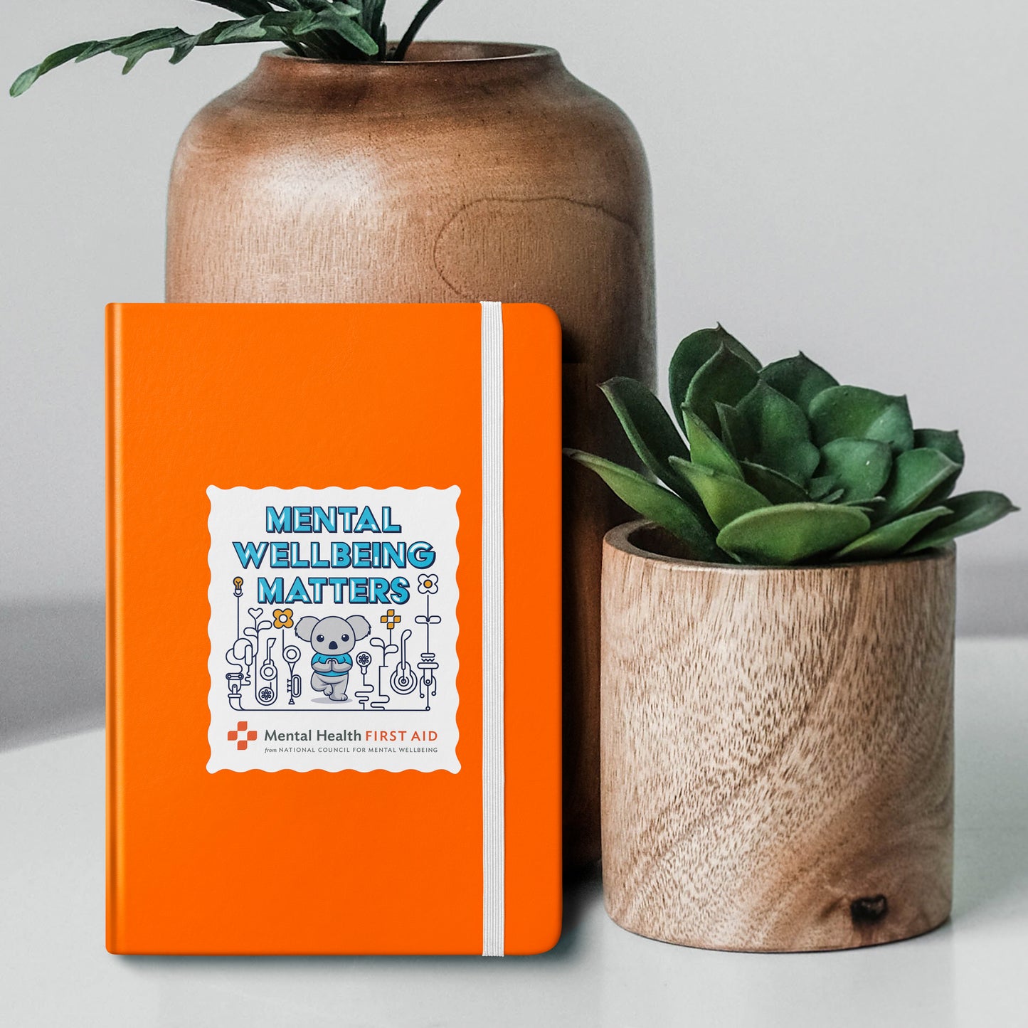 Orange notebook with 'Mental Wellbeing Matters' design, placed between two potted plants on a light background.