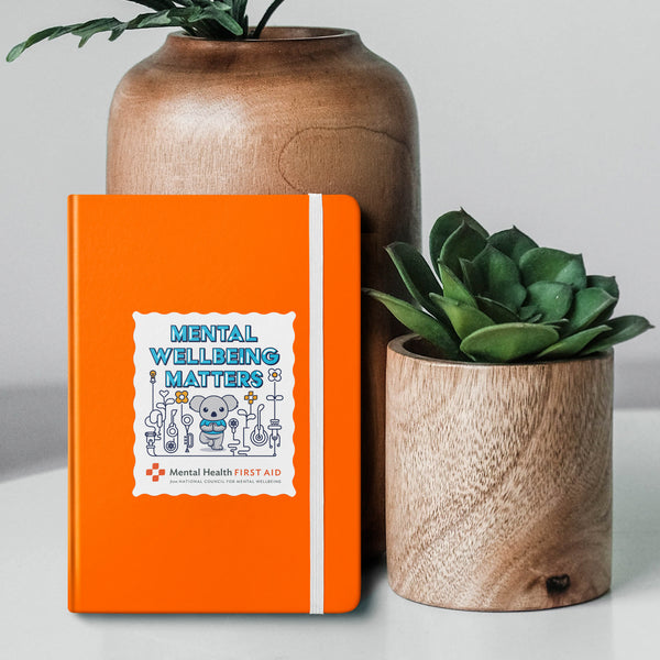 Orange notebook with 'Mental Wellbeing Matters' design, placed between two potted plants on a light background.