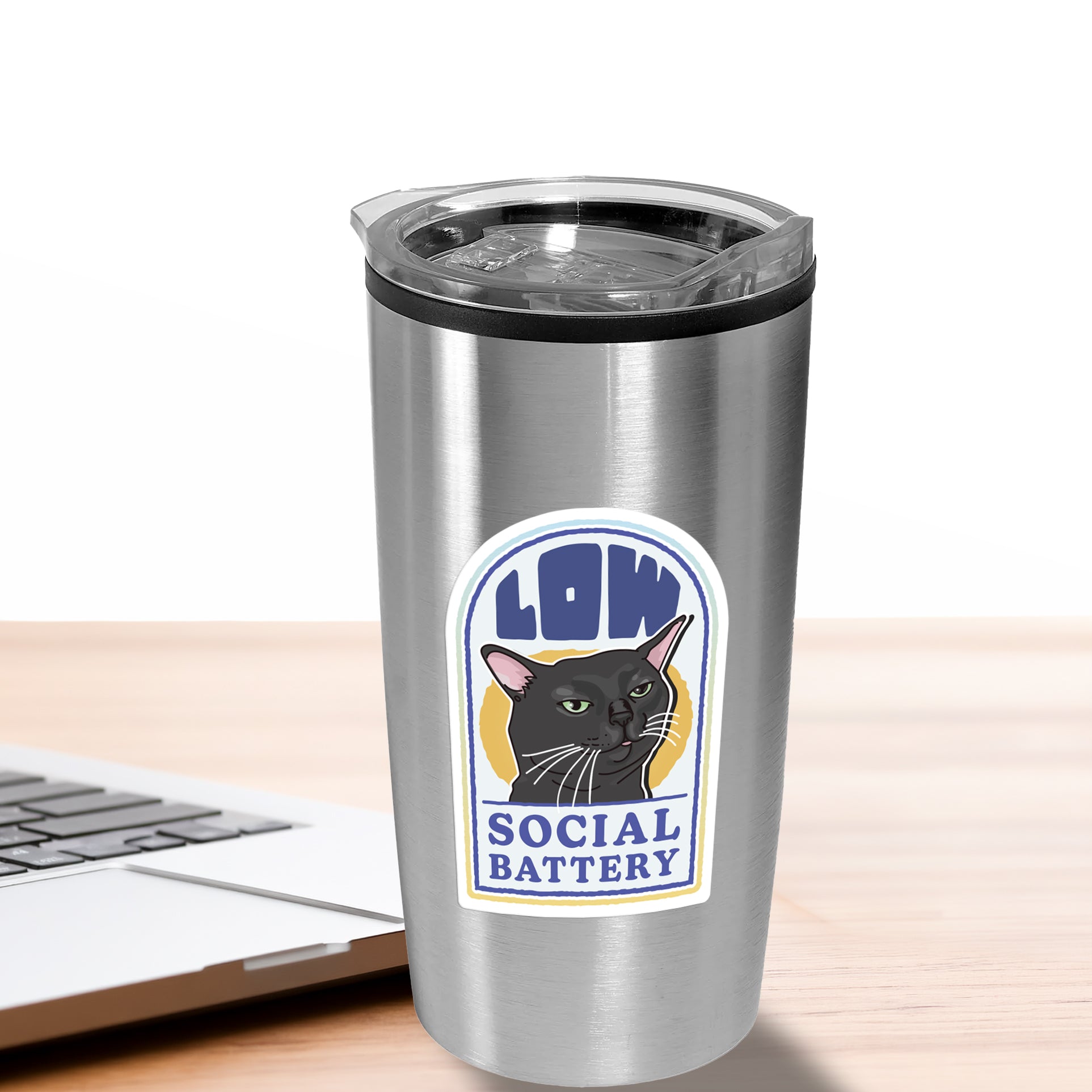Silver tumbler with a cat sticker labeled 'Low Social Battery' on a wooden surface with a laptop.