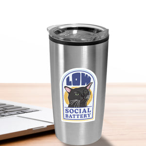 Silver tumbler with a cat sticker labeled 'Low Social Battery' on a wooden surface with a laptop.